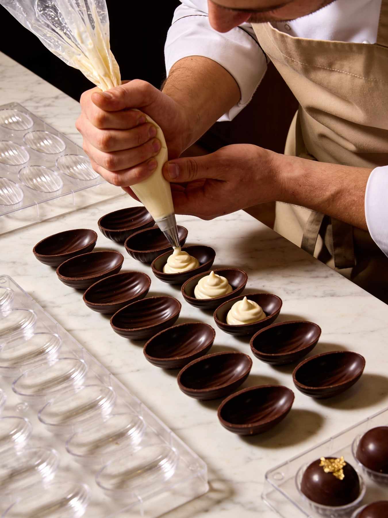 Hands piping ganache into rows of dark chocolate shells on a marble worktop.
