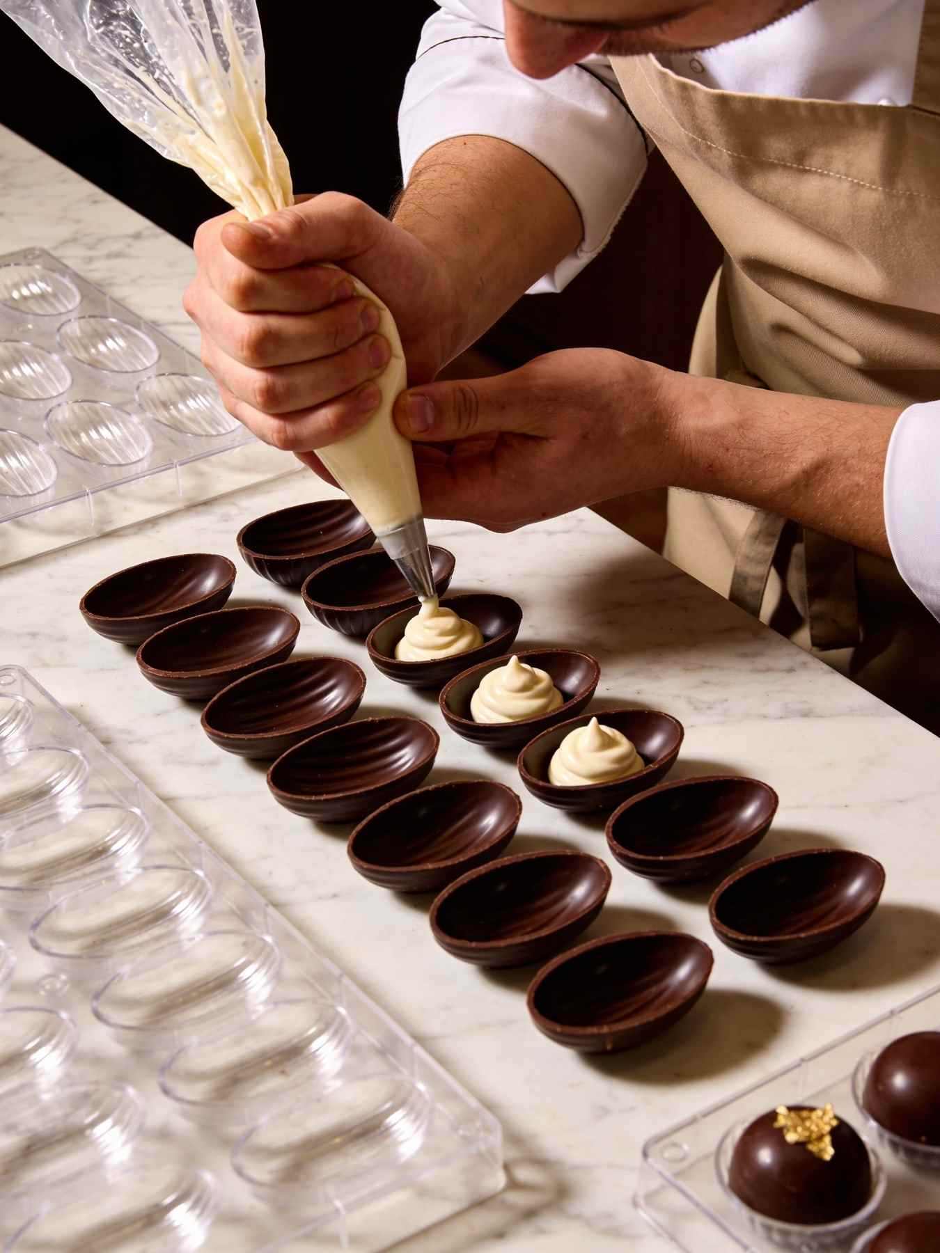 Hands piping ganache into rows of dark chocolate shells on a marble worktop.