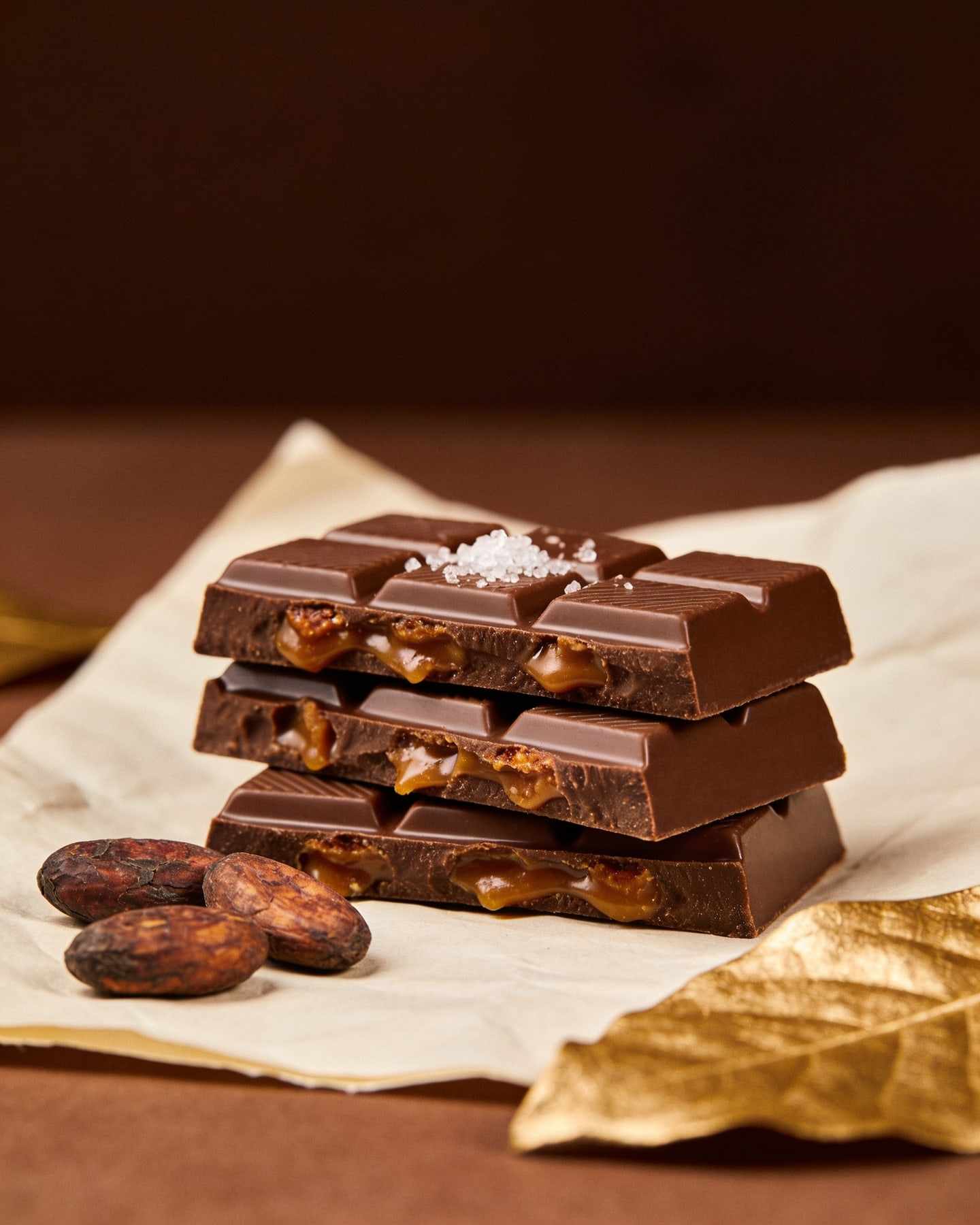 Stacked pieces of a creamy milk chocolate bar with caramel, cacao beans and fleur de sel on cream paper.