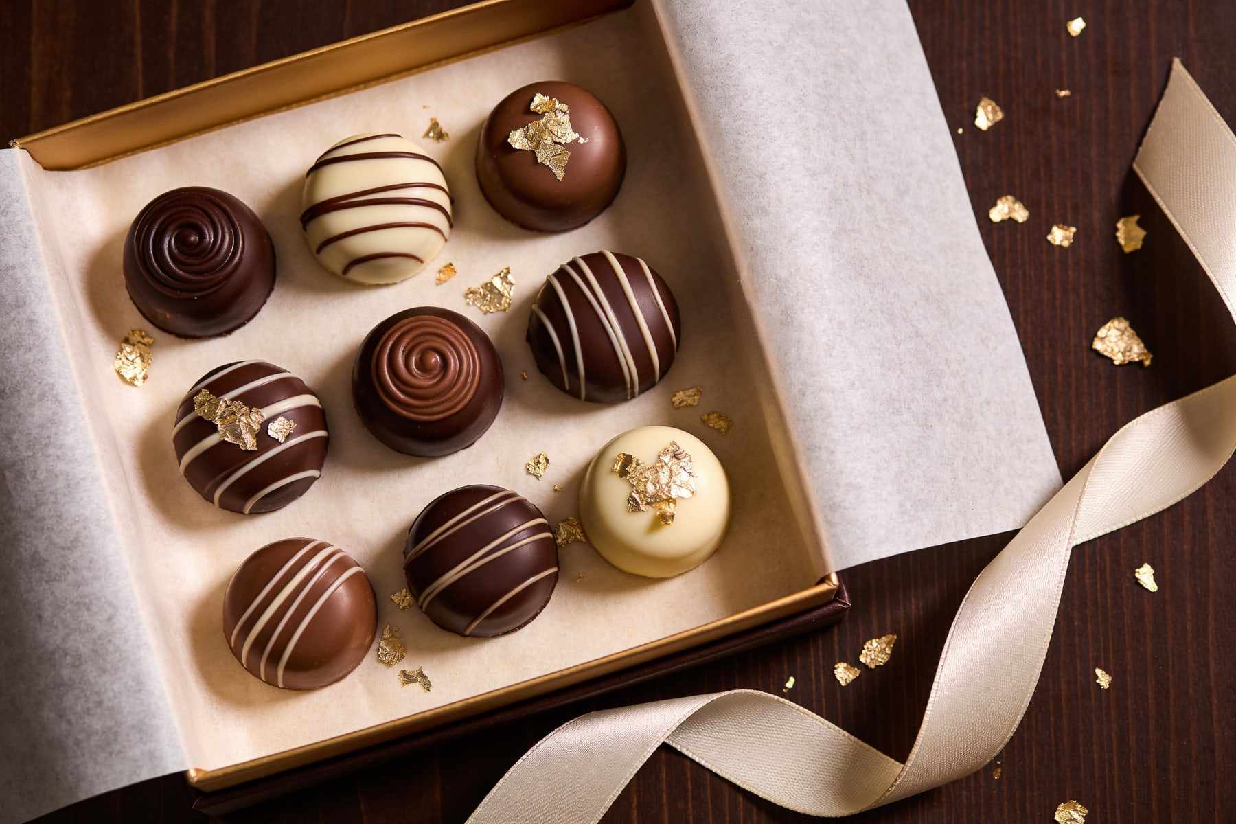An open gift box of nine hand-painted bonbons on cream tissue paper with a silk ribbon and gold leaf.