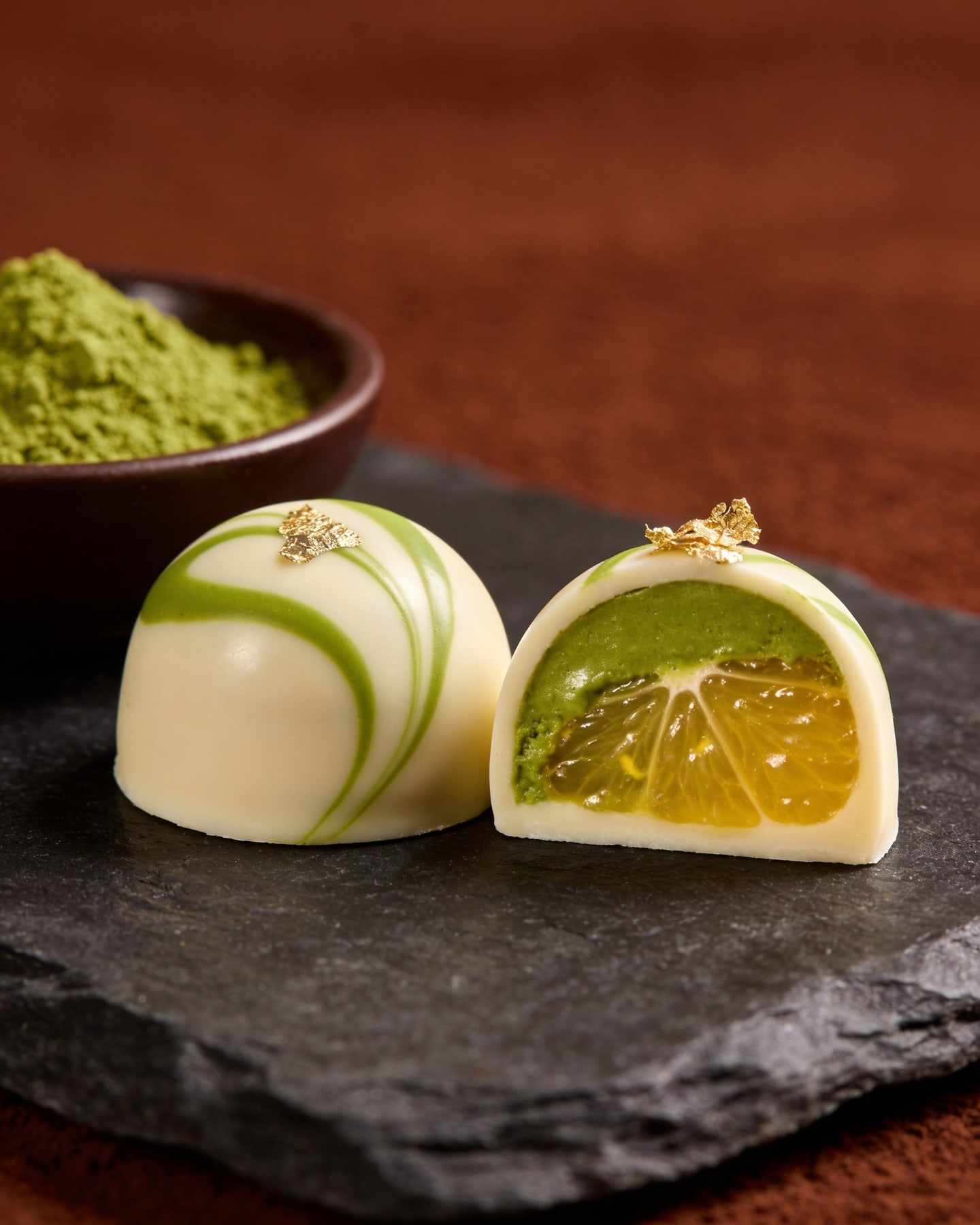 A white chocolate bonbon with pale green swirls, halved to show a matcha and yuzu ganache, matcha powder in a bowl.