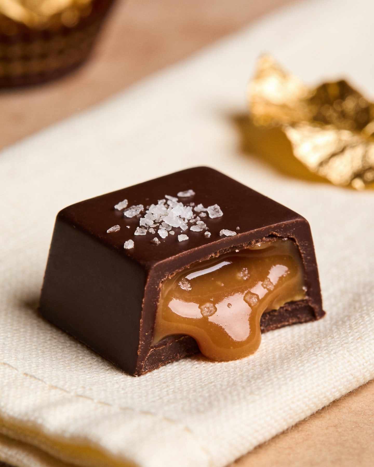 A square dark bonbon topped with sea salt flakes, halved with salted caramel flowing out, on cream linen.