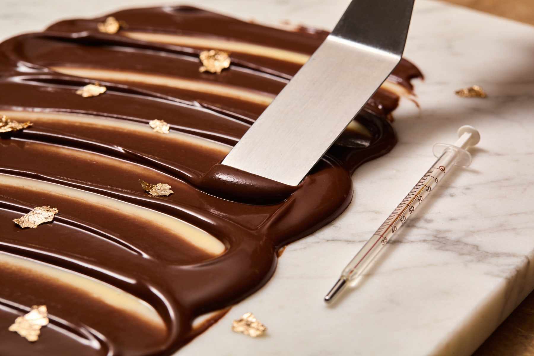Dark chocolate being tabled on a marble slab with a steel scraper, a thermometer beside it.